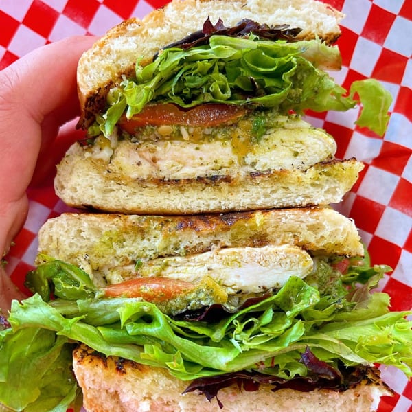 A hand holds a grilled chicken sandwich with pesto, lettuce, and tomato on toasted bread.