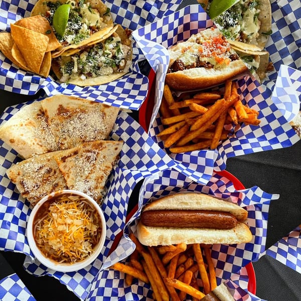 Assortment of Blacktop Grill street food: tacos, hot dogs, fries, quesadillas, and chili in checkered baskets.