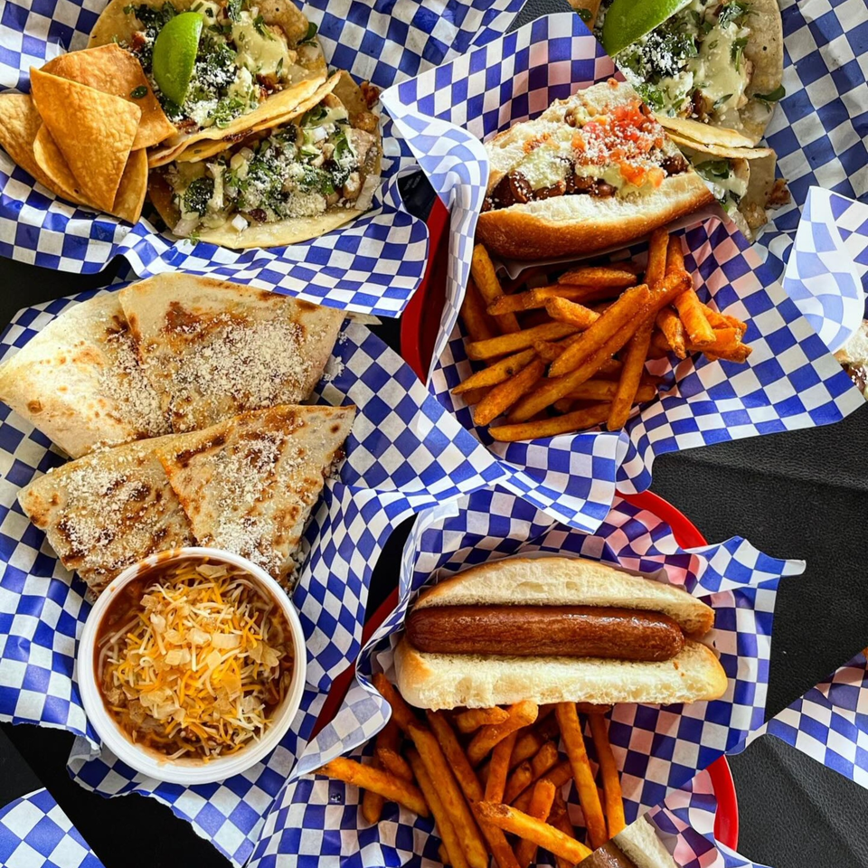 Assortment of Blacktop Grill street food: tacos, hot dogs, fries, quesadillas, and chili in checkered baskets.