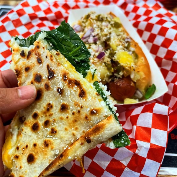 Hand holding a quesadilla with spinach and cheese, with a hot dog in the background.