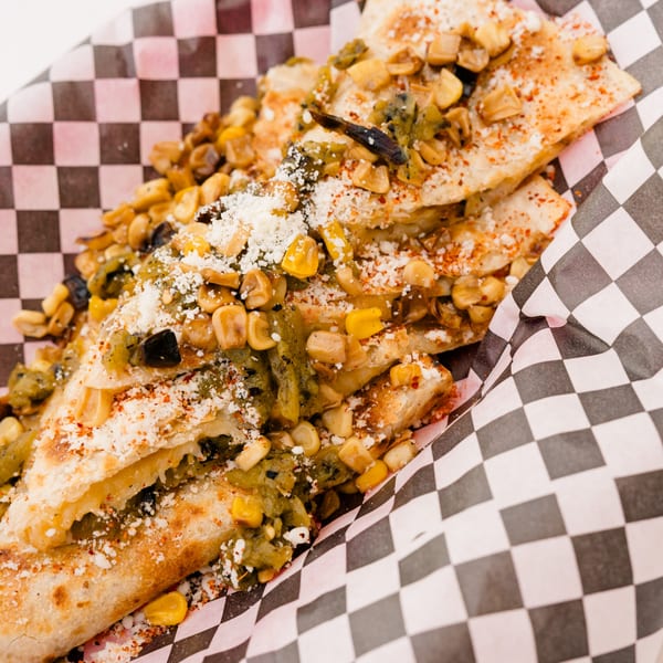 Quesadillas topped with corn, green chiles, cotija cheese, and chili powder in checkered paper.