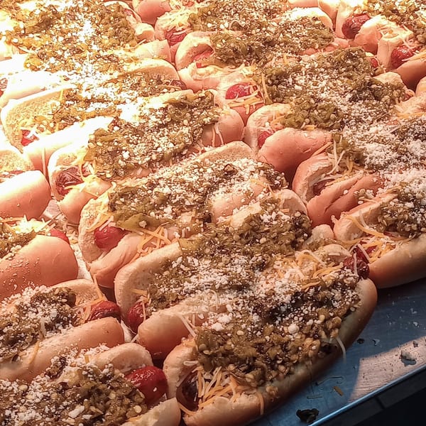 Numerous hot dogs in buns, topped with chili, shredded cheese, and white sprinkles, under bright lights.