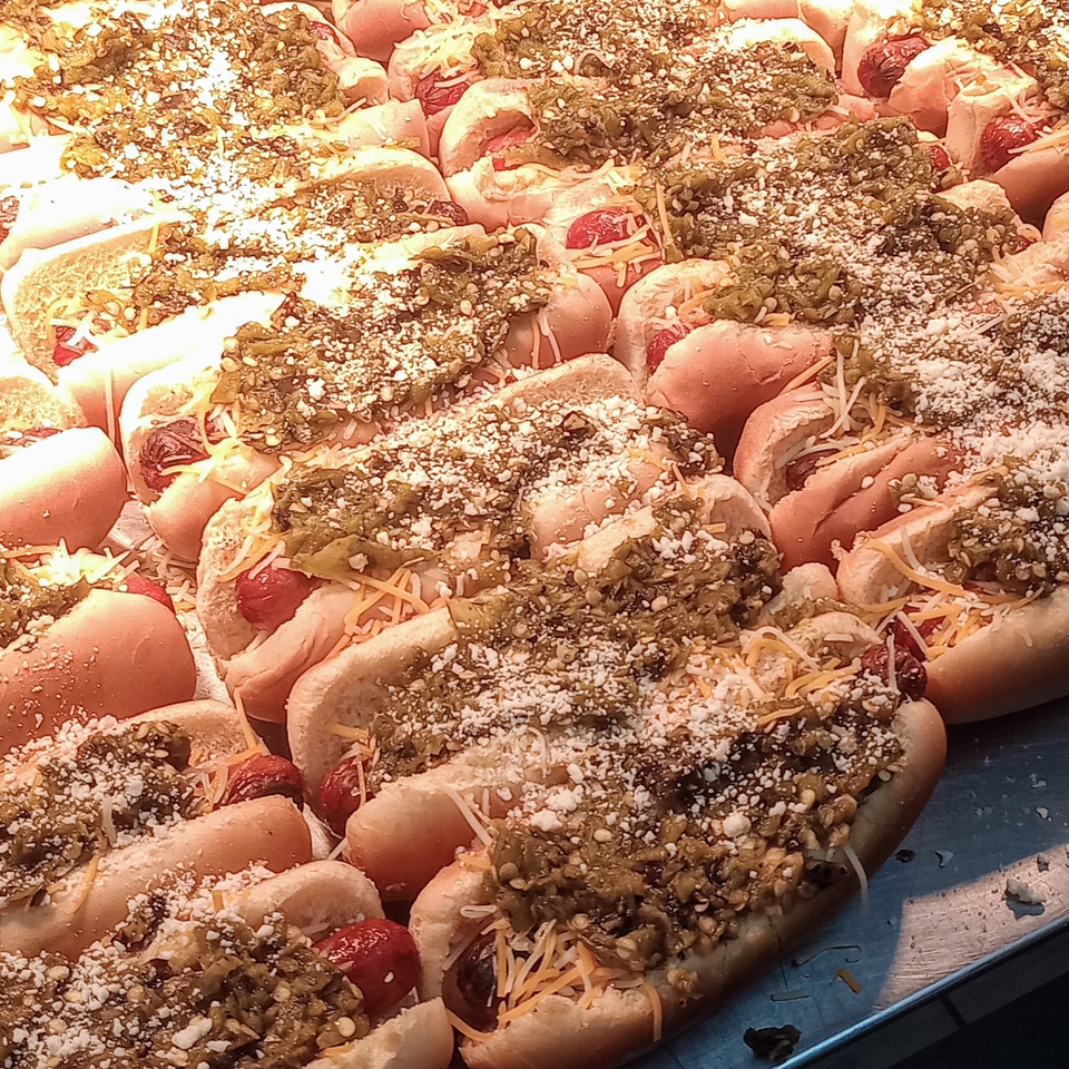Numerous hot dogs in buns, topped with chili, shredded cheese, and white sprinkles, under bright lights.