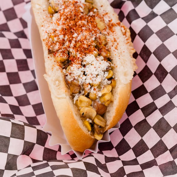 A close-up of a hot dog bun filled with corn, mushrooms, cheese, and red spice.