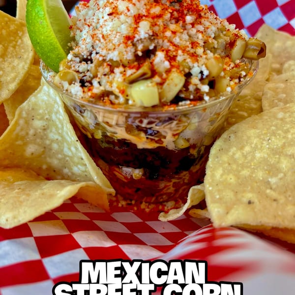 Mexican street corn in a cup with a lime wedge, surrounded by tortilla chips on a red and white checkered paper.