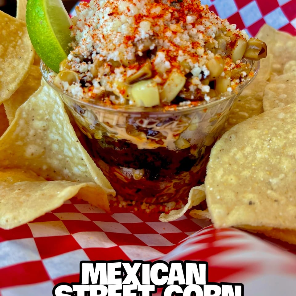 Mexican street corn in a cup with a lime wedge, surrounded by tortilla chips on a red and white checkered paper.