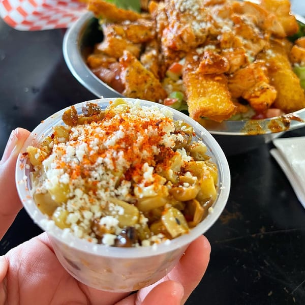 A hand holds a small cup of street corn with a large bowl of chicken and fries in the background.