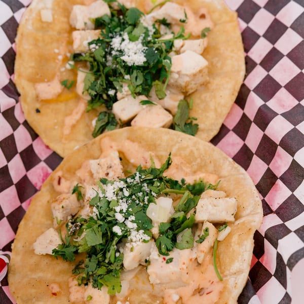 Two chicken tacos with cilantro, cheese, and sauce on a checkered paper liner.