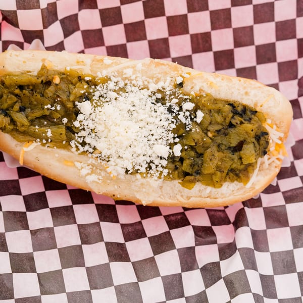 Hot dog in a bun topped with green chili, shredded cheese, and crumbled white cheese.