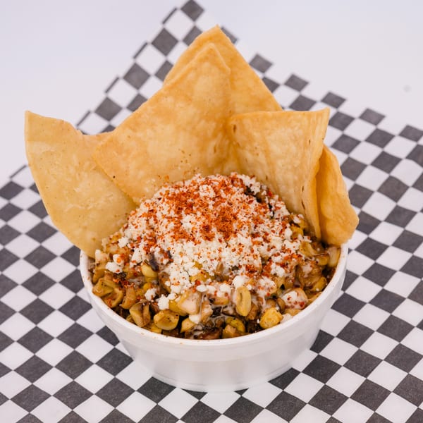 A white bowl of esquites with crumbled cheese, chili powder, and tortilla chips on a checkered napkin.