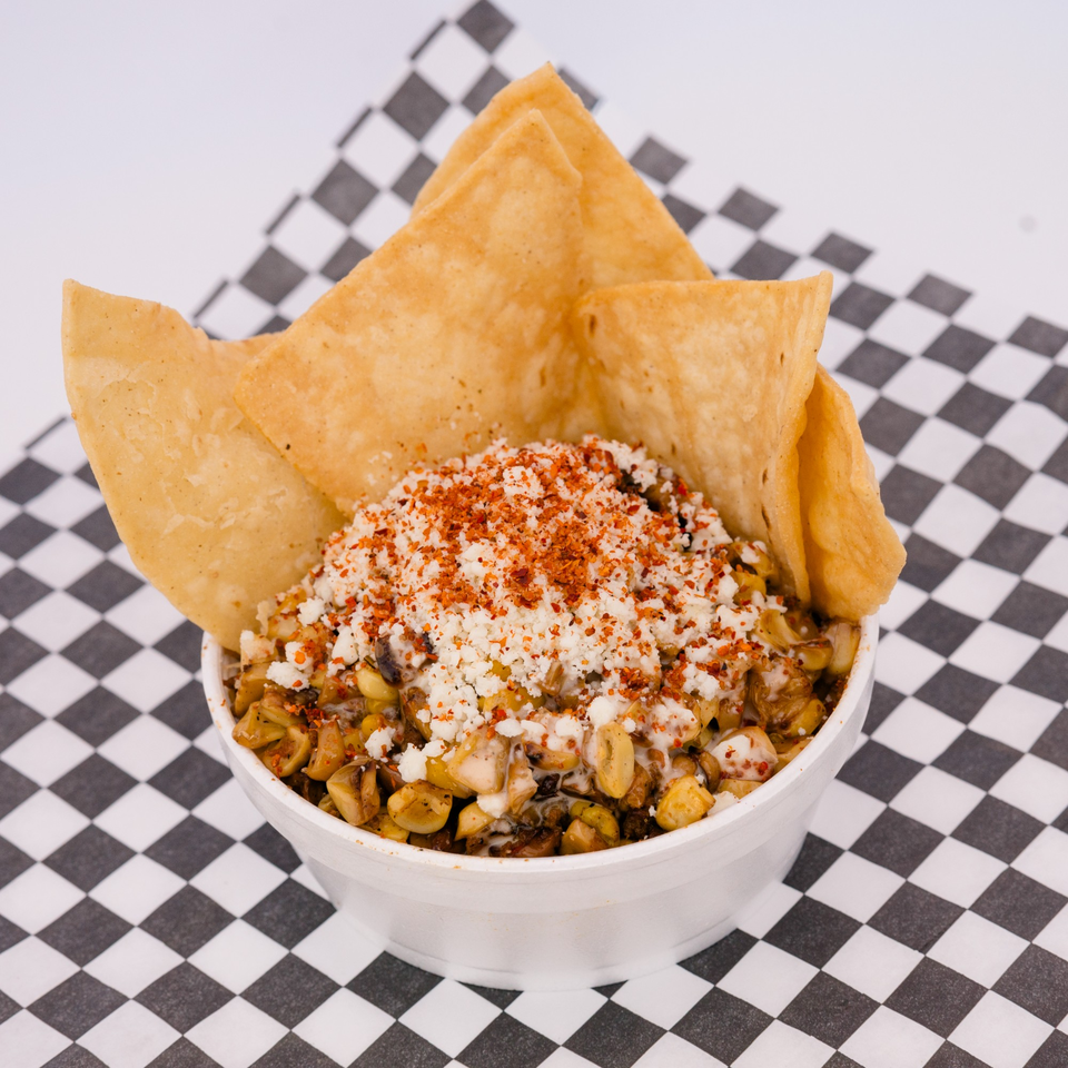 A white bowl of esquites with crumbled cheese, chili powder, and tortilla chips on a checkered napkin.