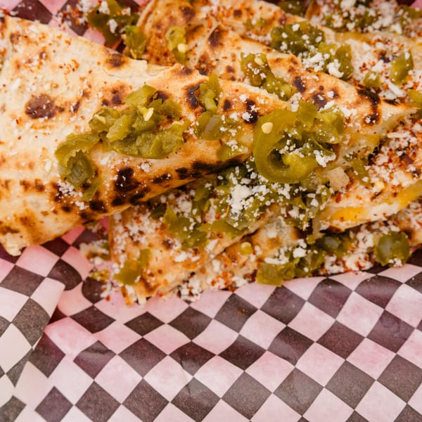 Close-up of quesadilla wedges topped with green chilies, cotija cheese, and red spices on checkered paper.