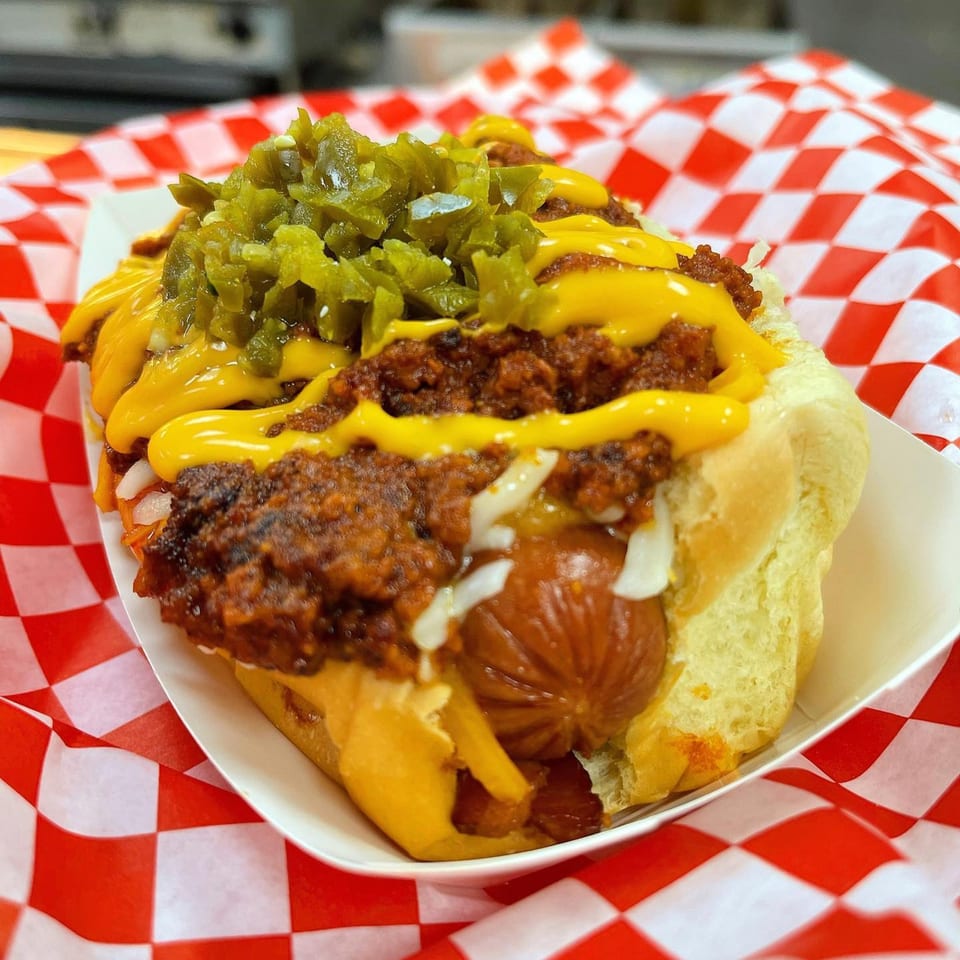 A chili cheese hot dog from The Blacktop Grill, topped with diced jalapeños, in a paper tray.